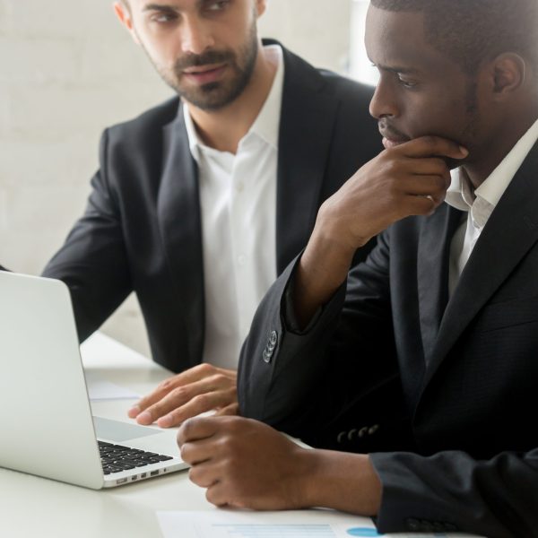 Serious black businessman thinking over business offer looking at laptop while caucasian partner explains deal details, successful african investor in suit considering investment listening to advisor