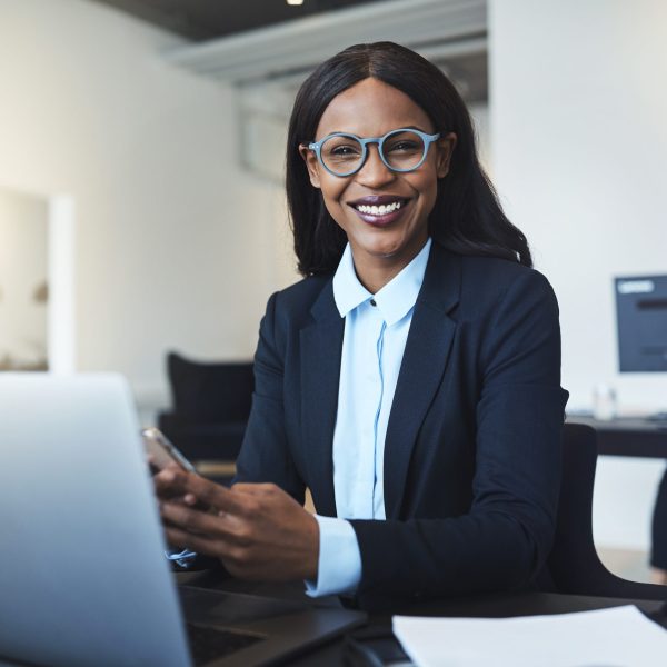 Smiling young African American businesswoman using a cellphone and working on a laptop while sitting at her desk in a bright modern office